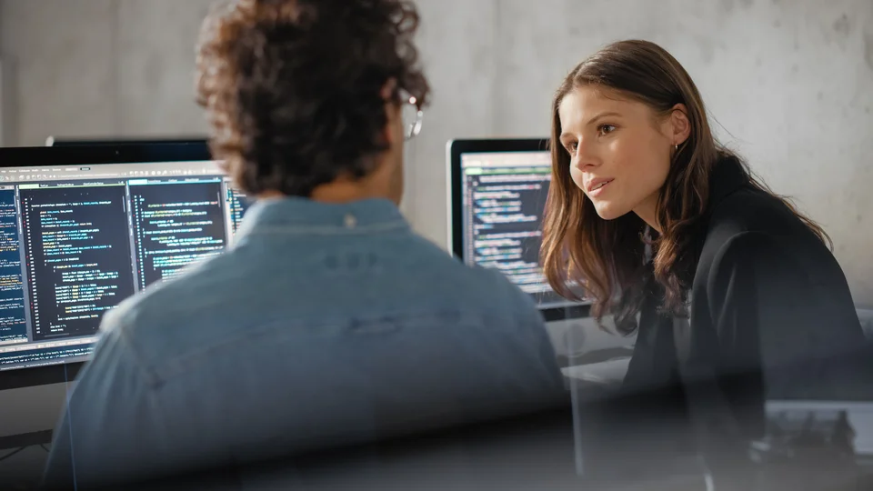 Eine Frau und ein Mann sitzen vor zwei Computern und diskutieren