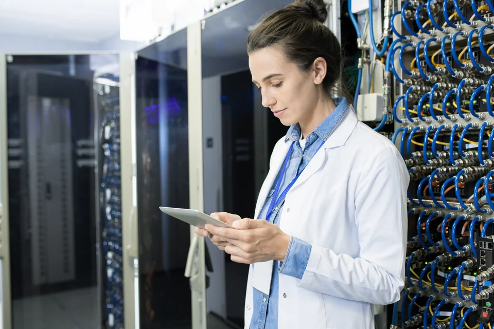 Frau im Laborkittel arbeitet in einem Serverraum, umgeben von technischen Geräten und Kabeln, fokussiert auf ihre Arbeit.
