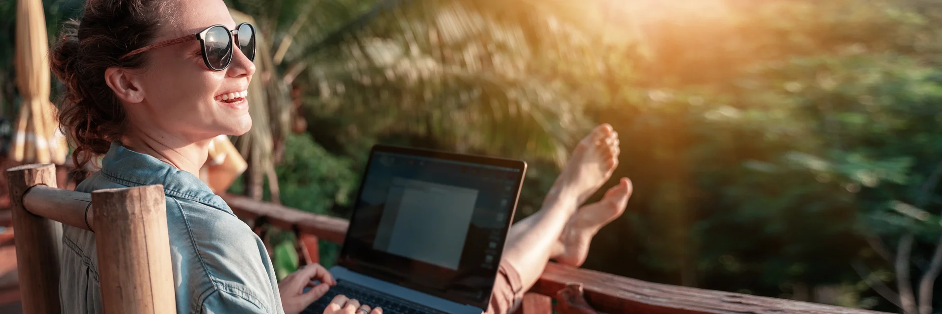 Eine lachende Frau sitzt auf einem Balkon mit Blick auf einen Wald udn arbeitet an einem Laptop.