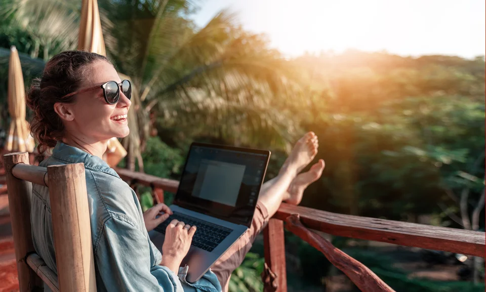 Eine lachende Frau sitzt auf einem Balkon mit Blick auf einen Wald udn arbeitet an einem Laptop.