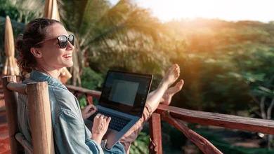 Eine lachende Frau sitzt auf einem Balkon mit Blick auf einen Wald udn arbeitet an einem Laptop.