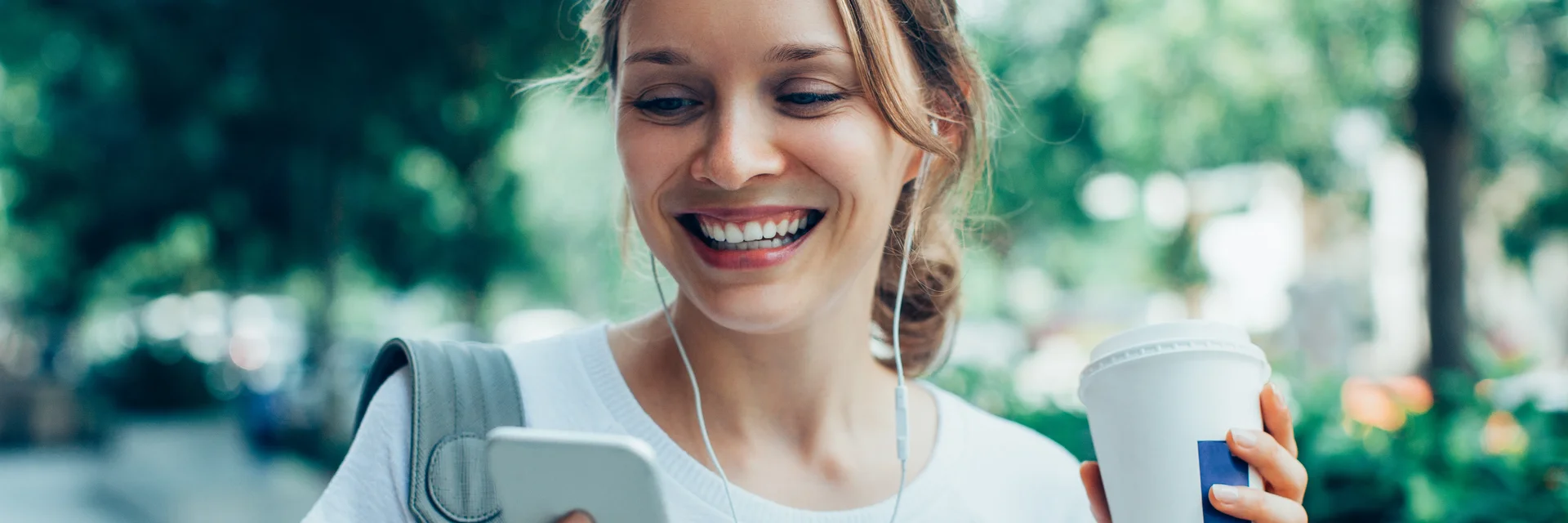 Eine Frau geht durch einen Park und schaut lächelnd auf ihr Handy in der rechten Hand. In der linken Hand hält sie einen Kaffee-Becher
