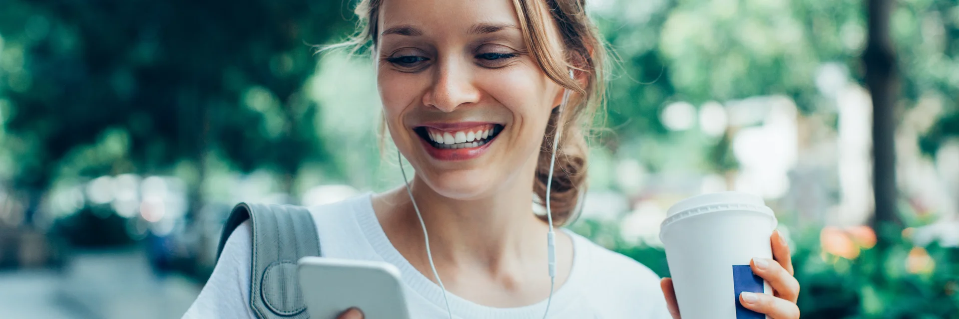 Eine Frau geht durch einen Park und schaut lächelnd auf ihr Handy in der rechten Hand. In der linken Hand hält sie einen Kaffee-Becher